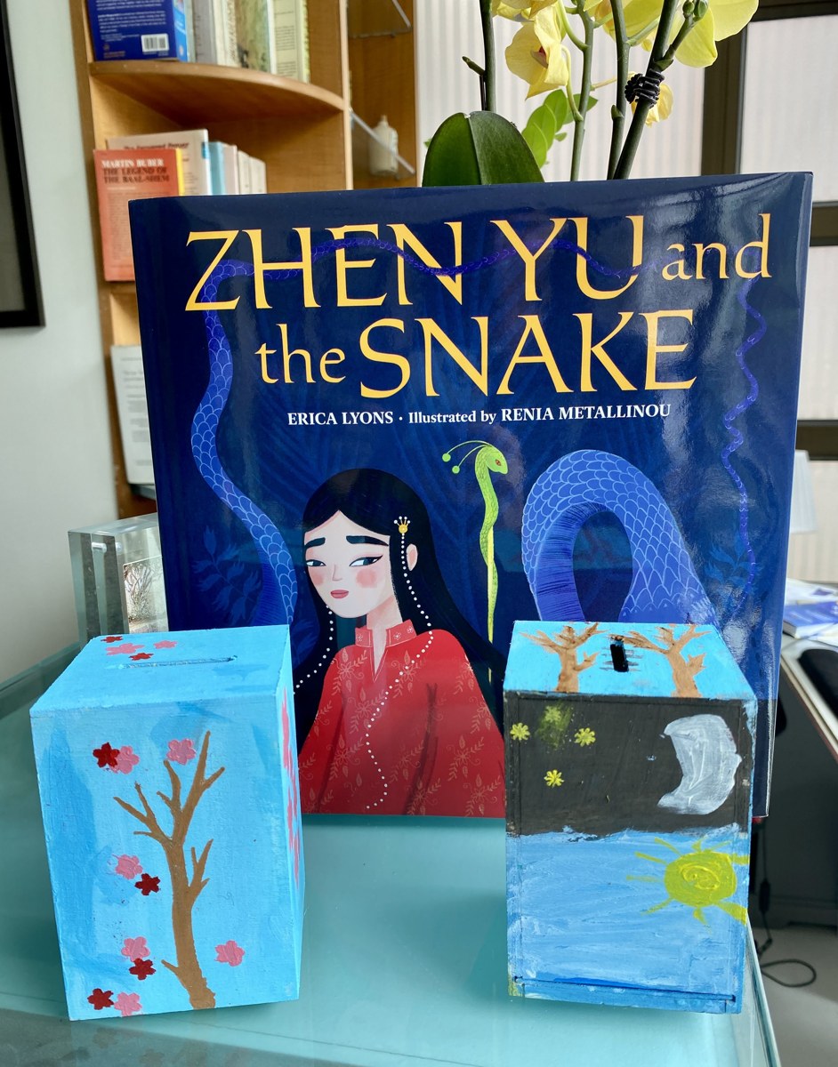 Finished tzedakah boxes inspired by Zhen Yu and the Snake, displayed with the book